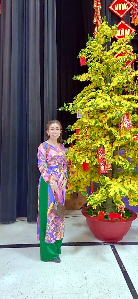 Woman in a colorful traditional dress posing beside a yellow flowering tree adorned with red envelopes on a stage with curtains in the background.