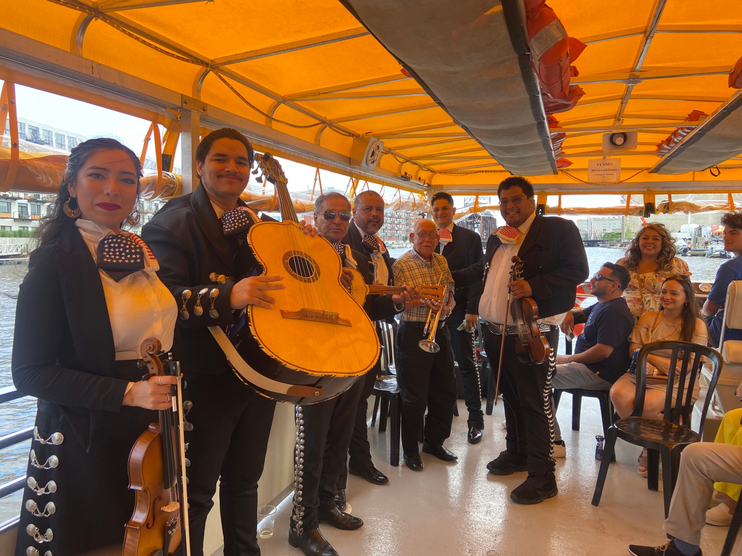 Mariachi musicians in black charro outfits performing on a boat with an orange canopy, guitar in foreground.