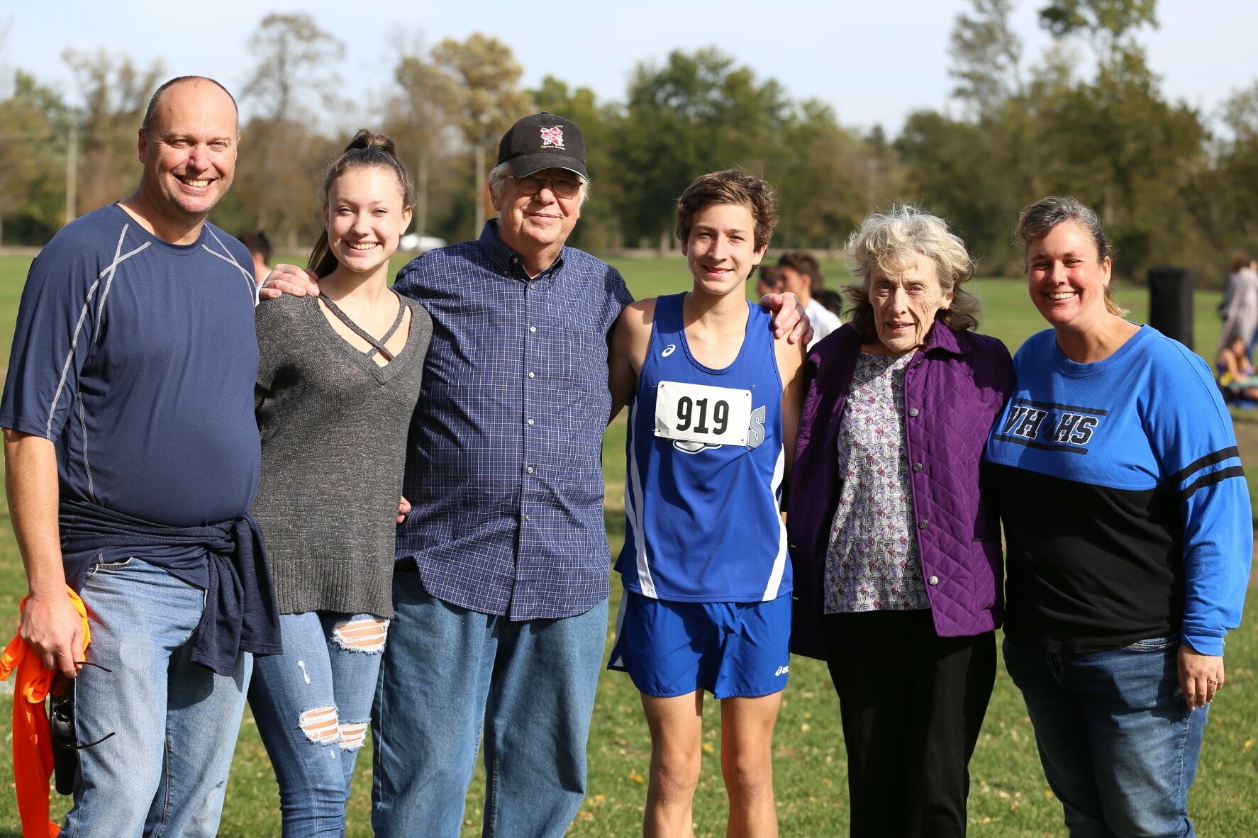 A diverse group of six people posing together outdoors on a grassy field after a race, smiling at the camera.