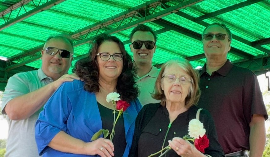 Five adults posing together under a green-roofed pavilion; two women in front hold red and white roses.