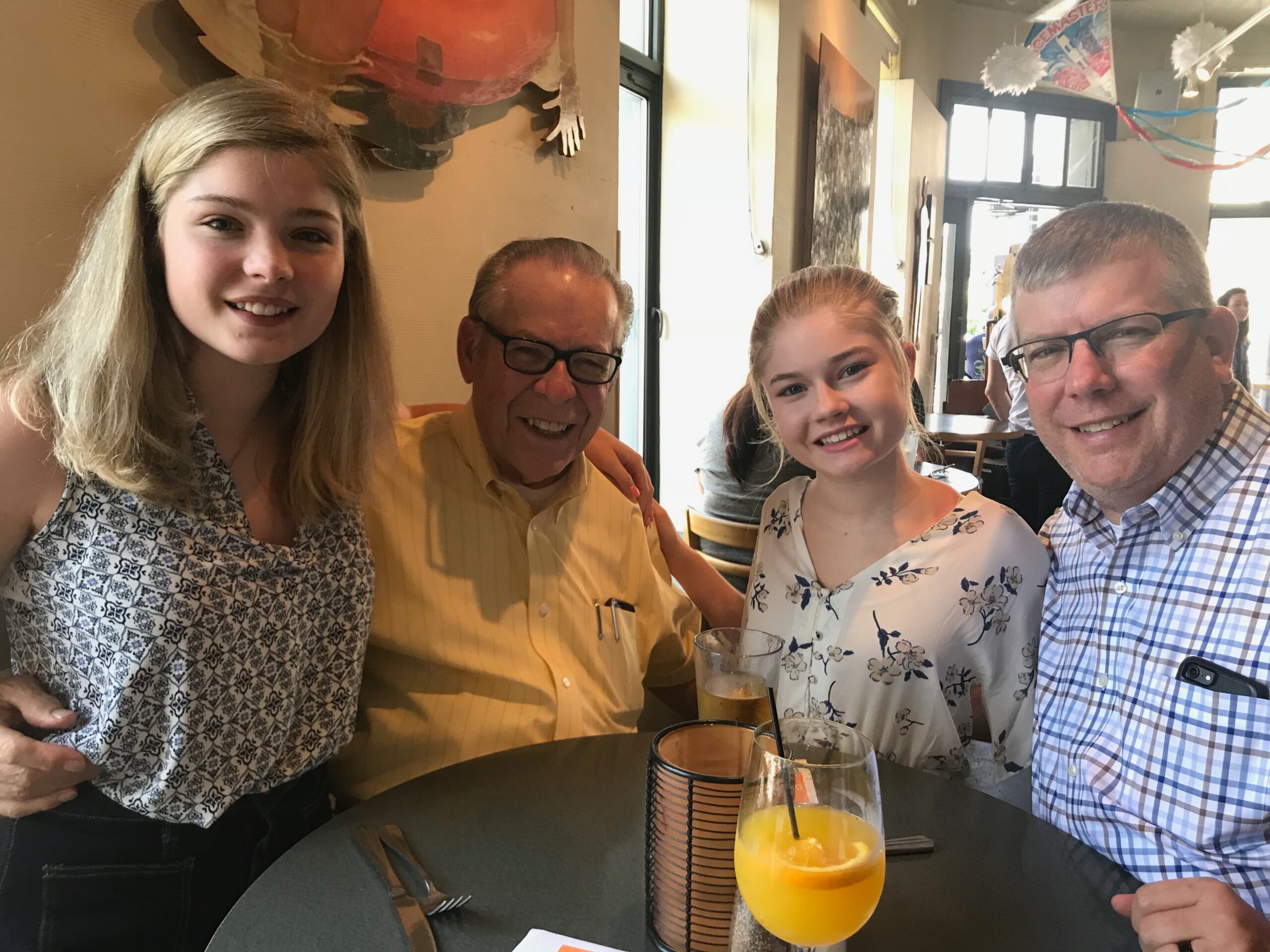 Four people smiling at a restaurant table, taking a group selfie with drinks in front of them.
