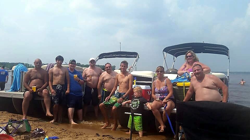 Group of adults and a child posing on a sandy beach in front of pontoon boats, some holding drinks and wearing swimwear.