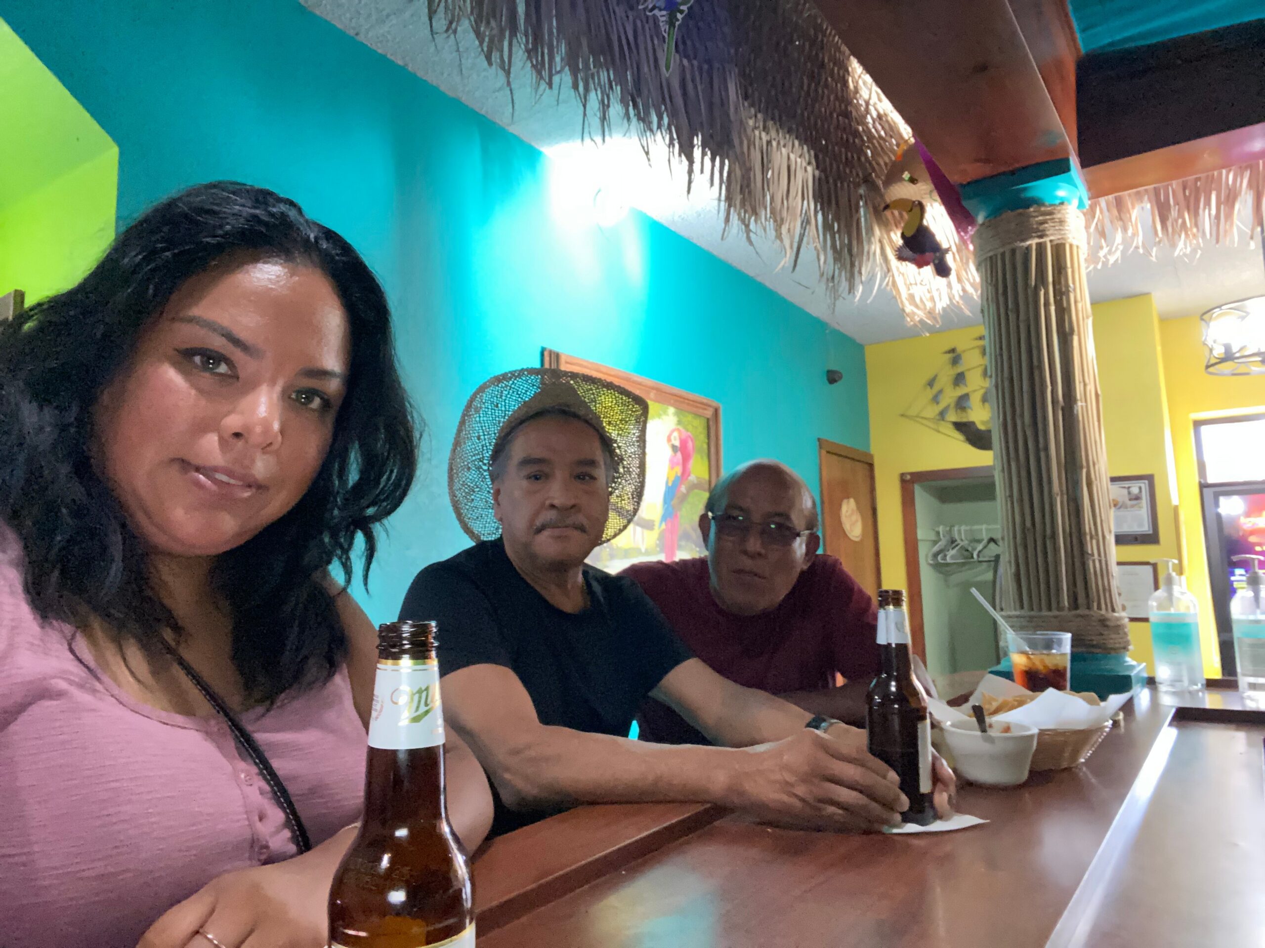 People sit at a tropical-themed bar counter with bottles, colorful turquoise and yellow walls, and thatch decor overhead.