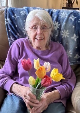 Smiling elderly woman with white hair and glasses, wearing a purple shirt, holding a glass vase of multicolored tulips.