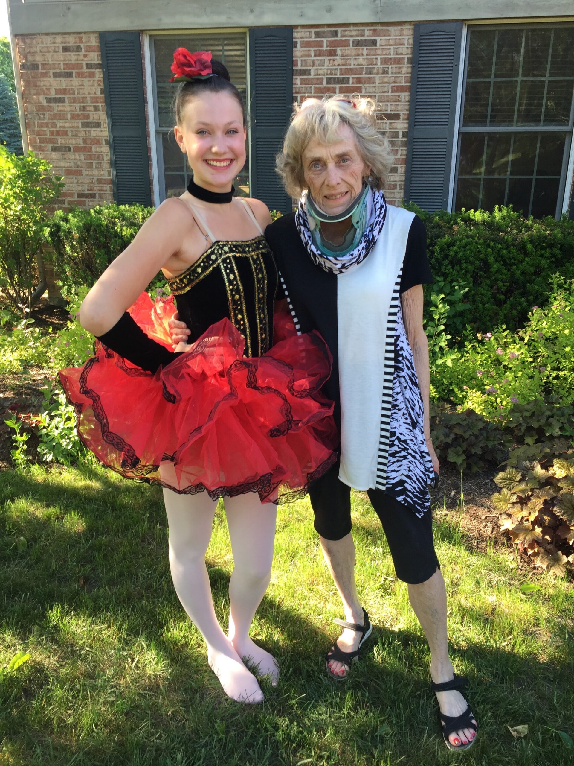 Smiling young ballerina in a red tutu and black bodice poses with an older person in a scarf outside a brick house.