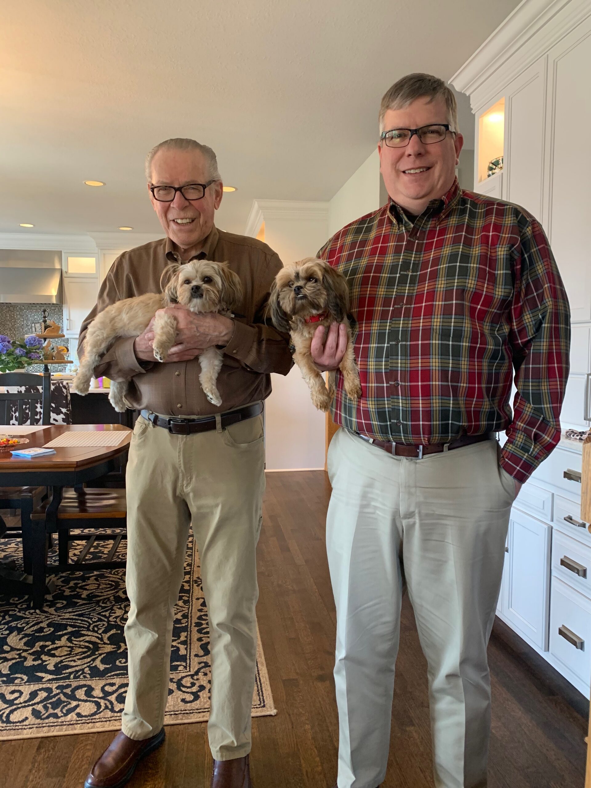 Two adults stand in a bright kitchen, each holding a small fluffy dog and smiling at the camera.