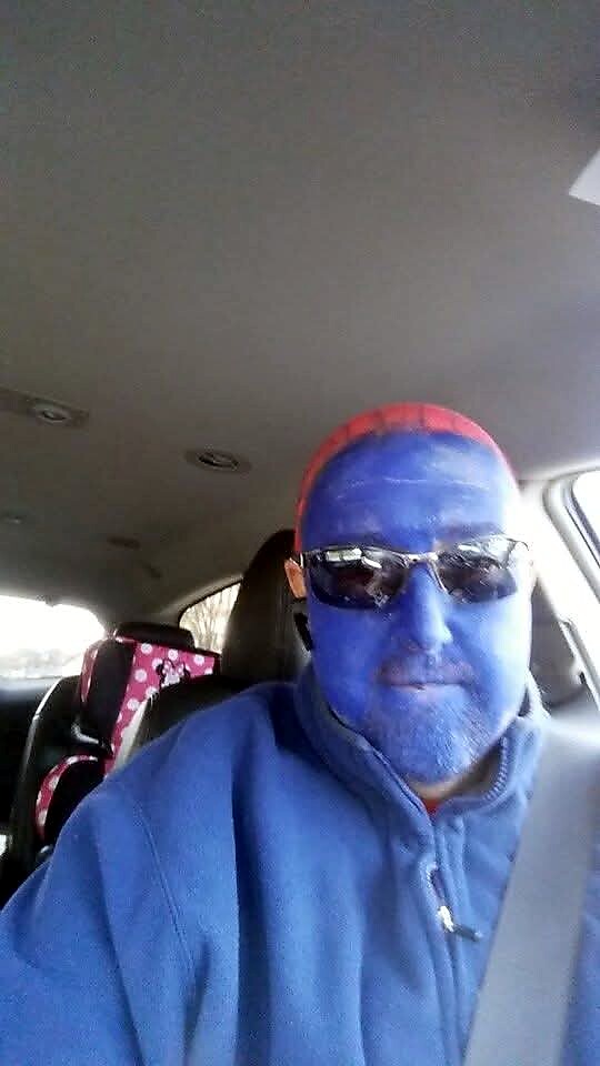 Person in a car wearing blue face paint, red Spider-Man cap, sunglasses, and a blue zip‑up jacket, taking a selfie with a seatbelt on.