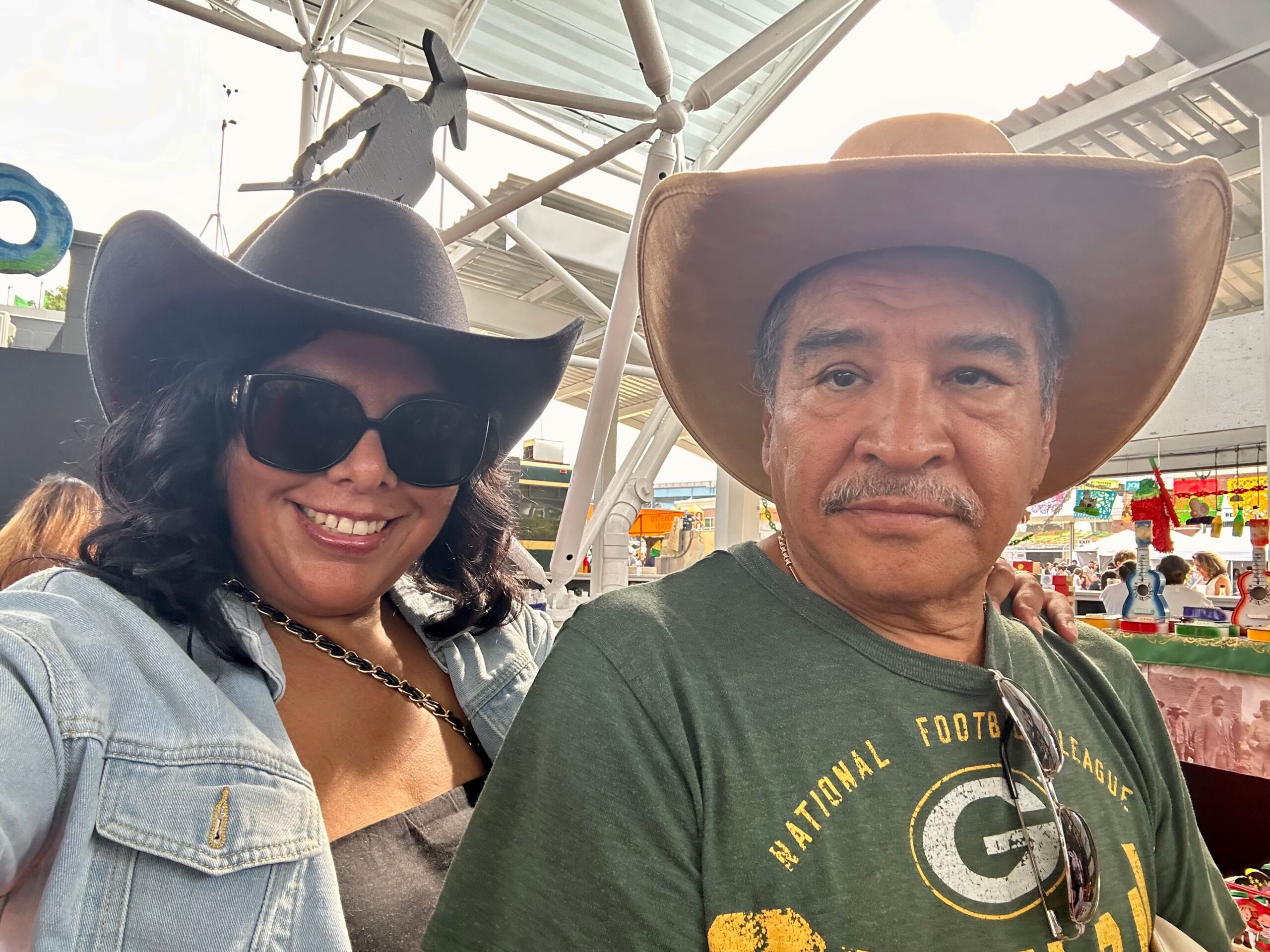 A smiling woman wearing a black cowboy hat and sunglasses next to a man with a tan wide-brimmed hat taking a selfie at an outdoor market.
