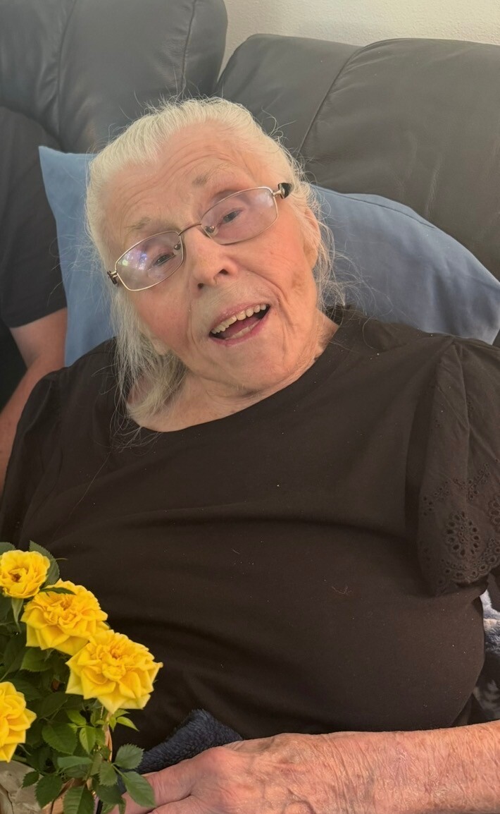 Elderly woman with white hair and glasses sitting on a sofa, wearing a black shirt, holding a bouquet of yellow roses.