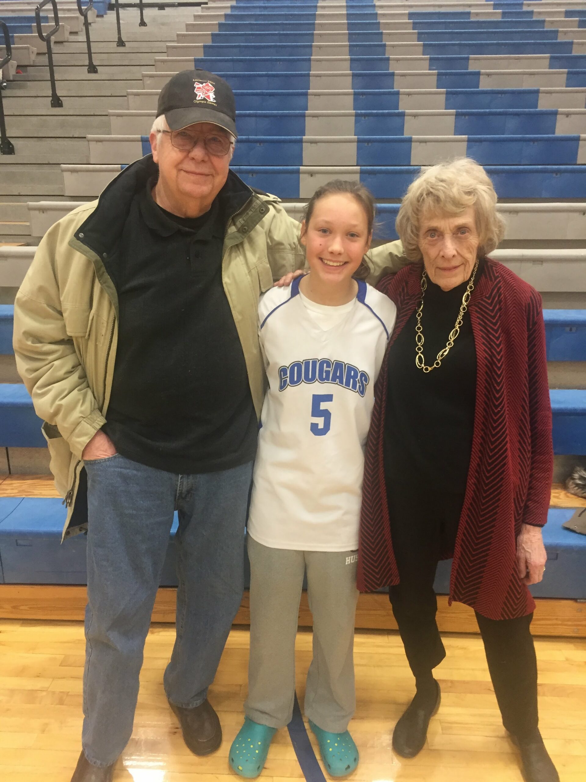 Three people posing together in a gym bleacher area: an older man in a cap and beige jacket, a smiling girl in a white and blue 'Cougars' jersey, and an older woman in a red cardigan and gold necklace.