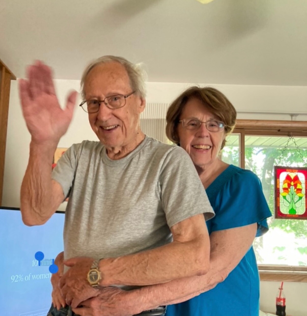 Elderly man in a gray t-shirt waves at the camera while an older woman in blue stands behind him, both smiling indoors.