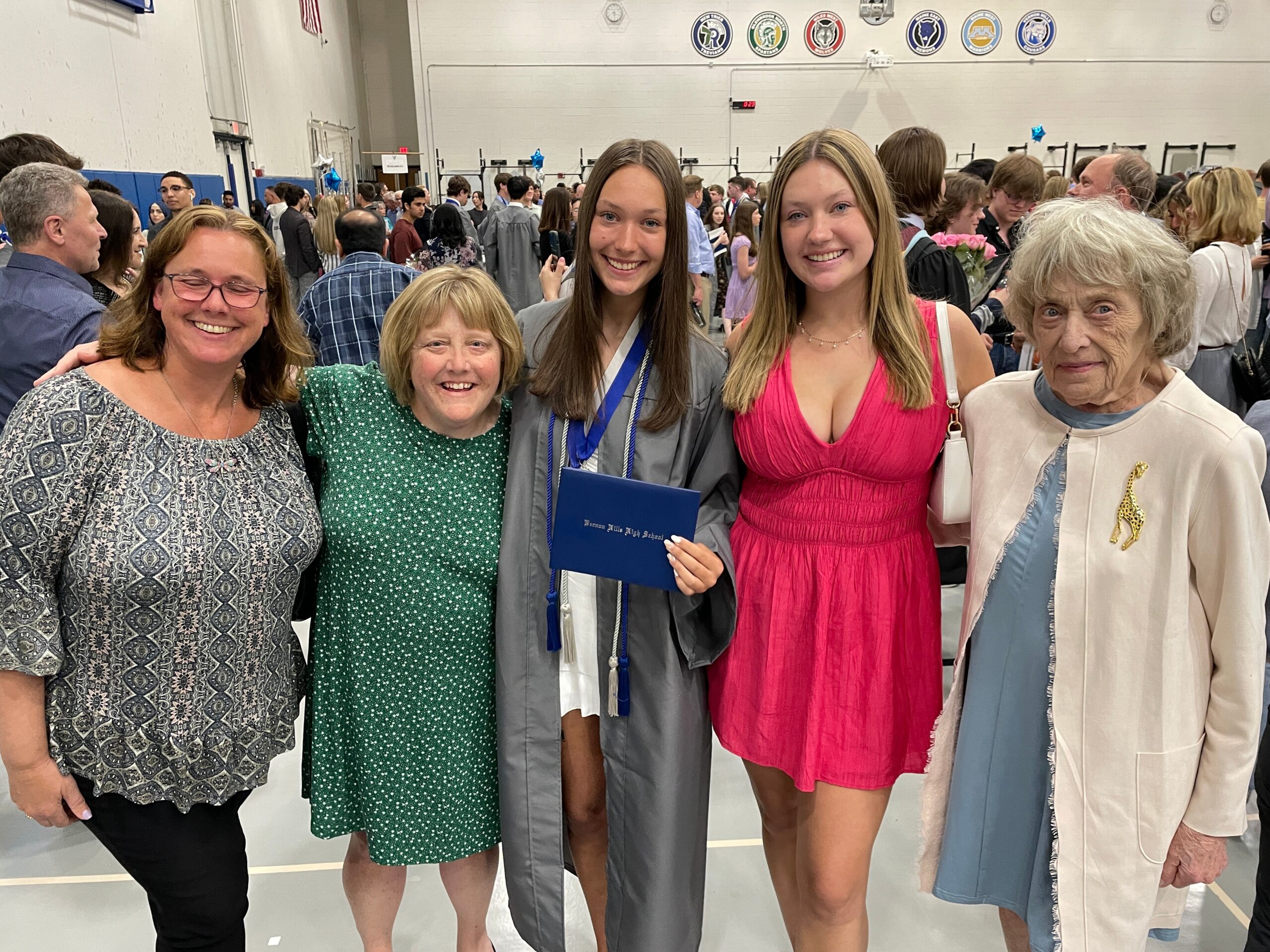 Group of graduates and family posing for a photo at a school graduation ceremony in a gymnasium filled with people in the background.