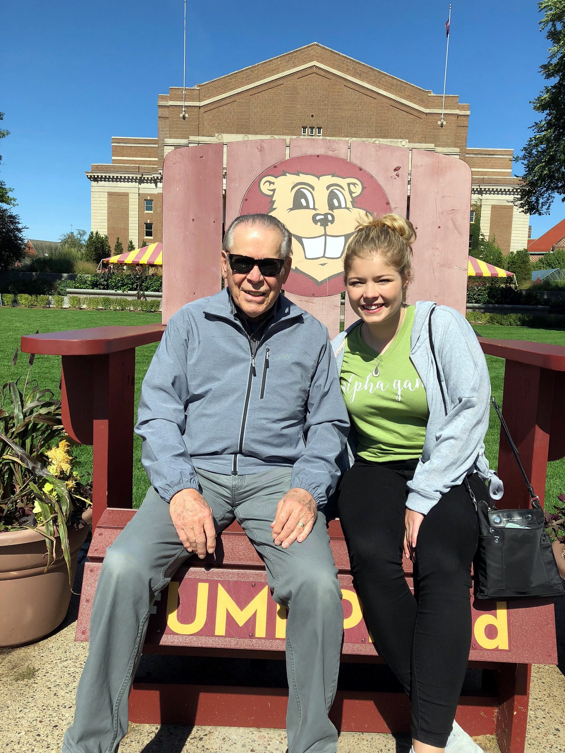 Two people sitting on a large wooden bench with a cartoon beaver sign behind them, smiling at the camera on a sunny day.