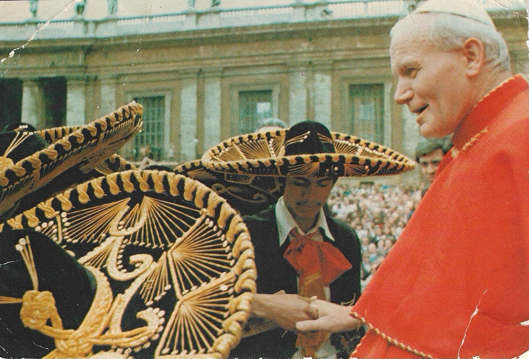 Cleric in red vestments smiles as large ornate sombreros are displayed in a public square with a crowd in the background.