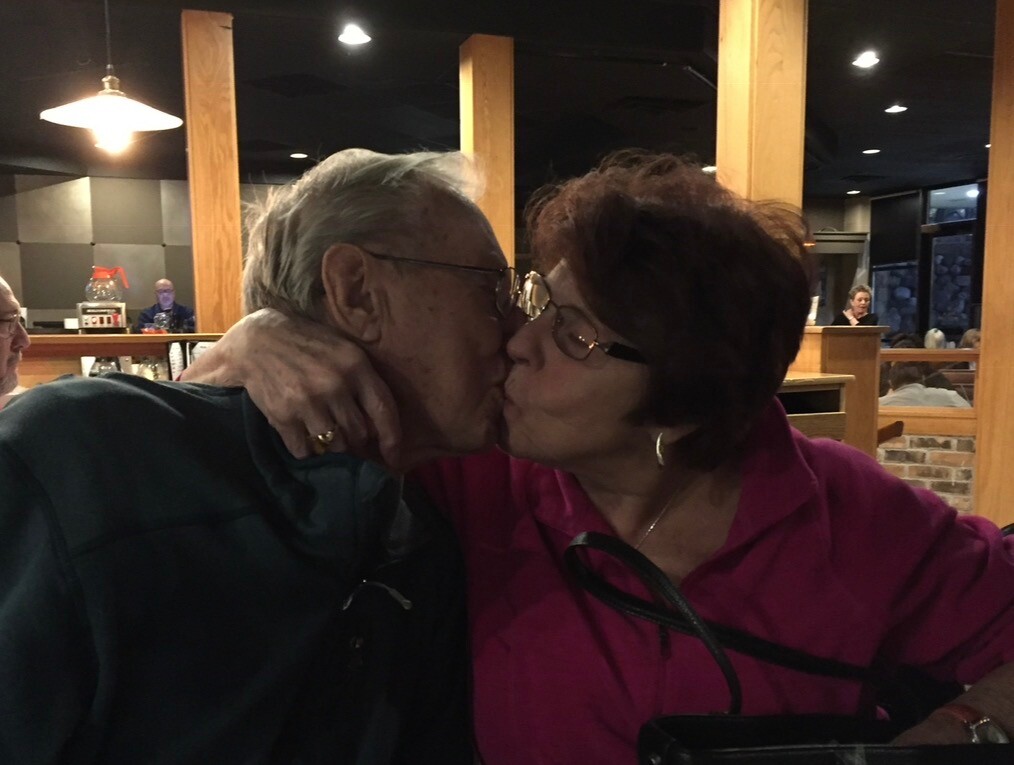 An elderly couple kissing at a restaurant table, the man in a dark shirt and the woman in a magenta jacket, with warm dining-room lighting behind them (close-up).