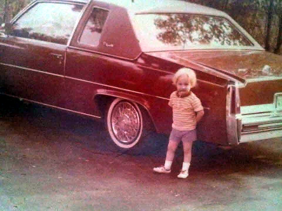 A toddler in a striped shirt and shorts stands beside a large vintage red car parked outdoors.