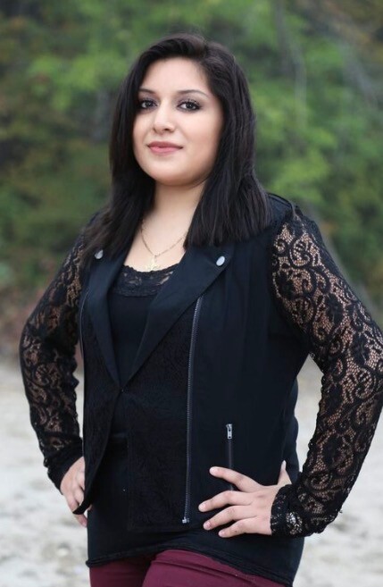 Portrait of a woman standing outdoors with hands on hips, wearing a black lace-sleeve top and vest.