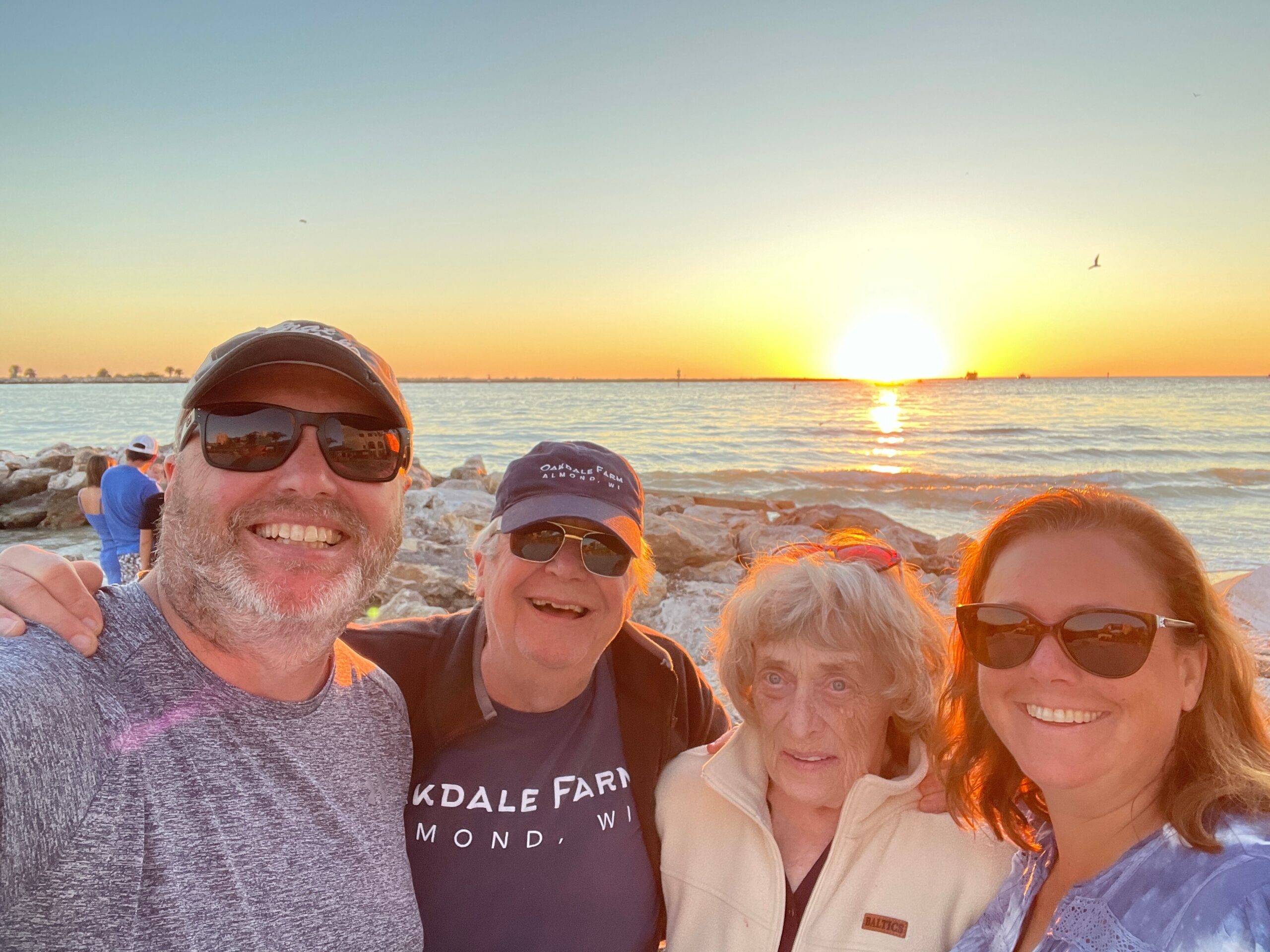 Group selfie at the beach during sunset with four people smiling up close to the camera, rocky shoreline in the foreground and calm sea behind them, warm orange sky.