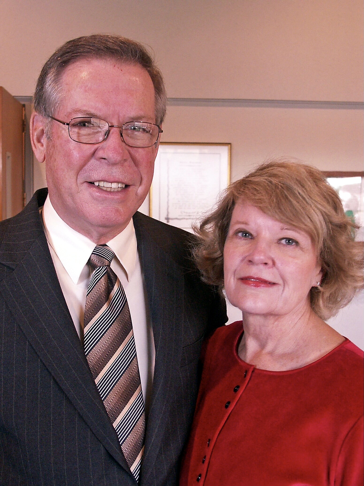 Smiling man in a dark pinstriped suit and striped tie standing next to a woman in a red blouse indoors.