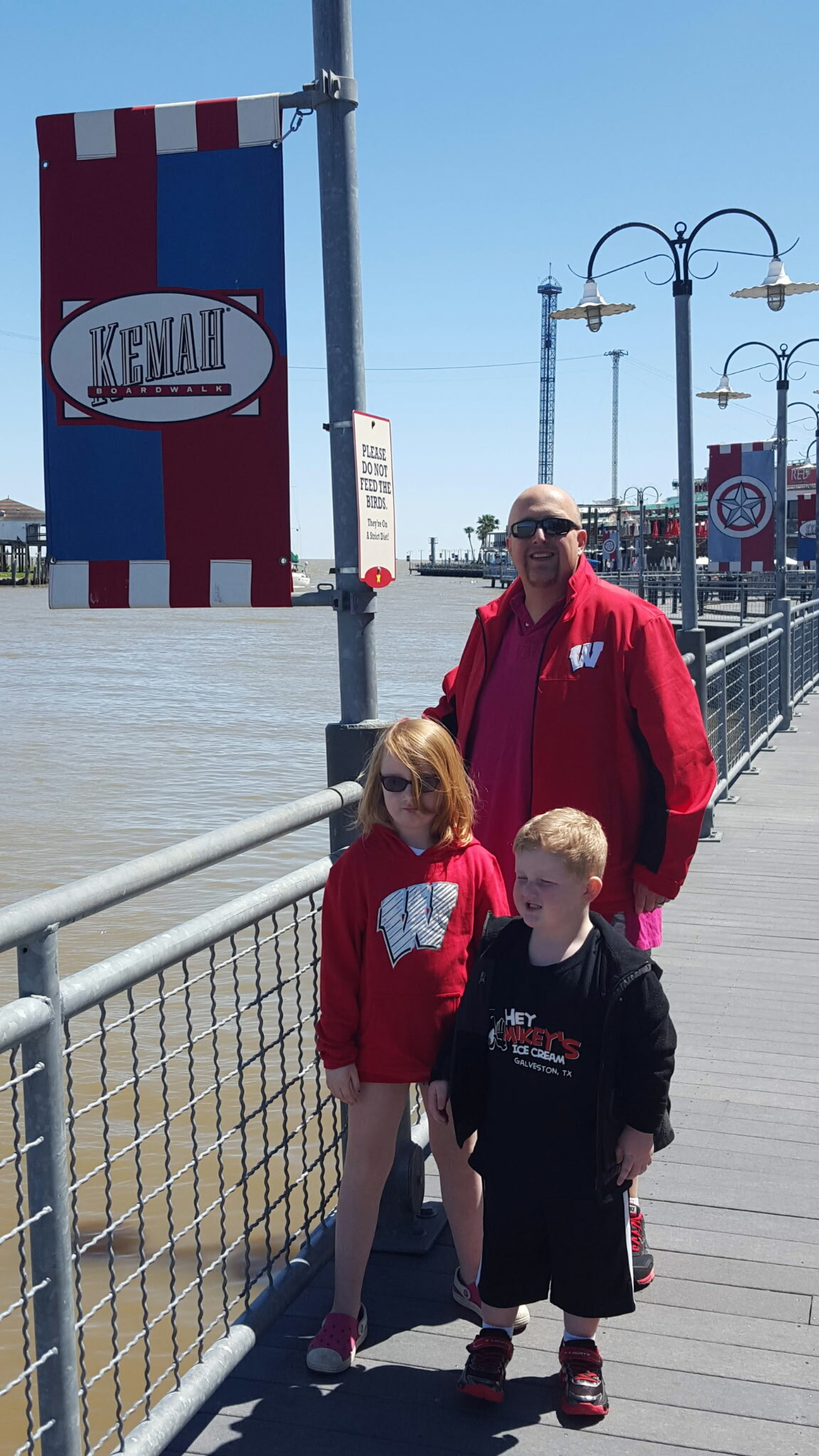 Man and two children stand on a sunny pier beside the water, wearing red jackets, with a Kemah Boardwalk banner nearby.