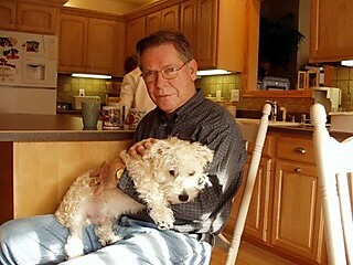 Older man in a plaid shirt sitting in a kitchen, holding a small fluffy white dog on his lap.