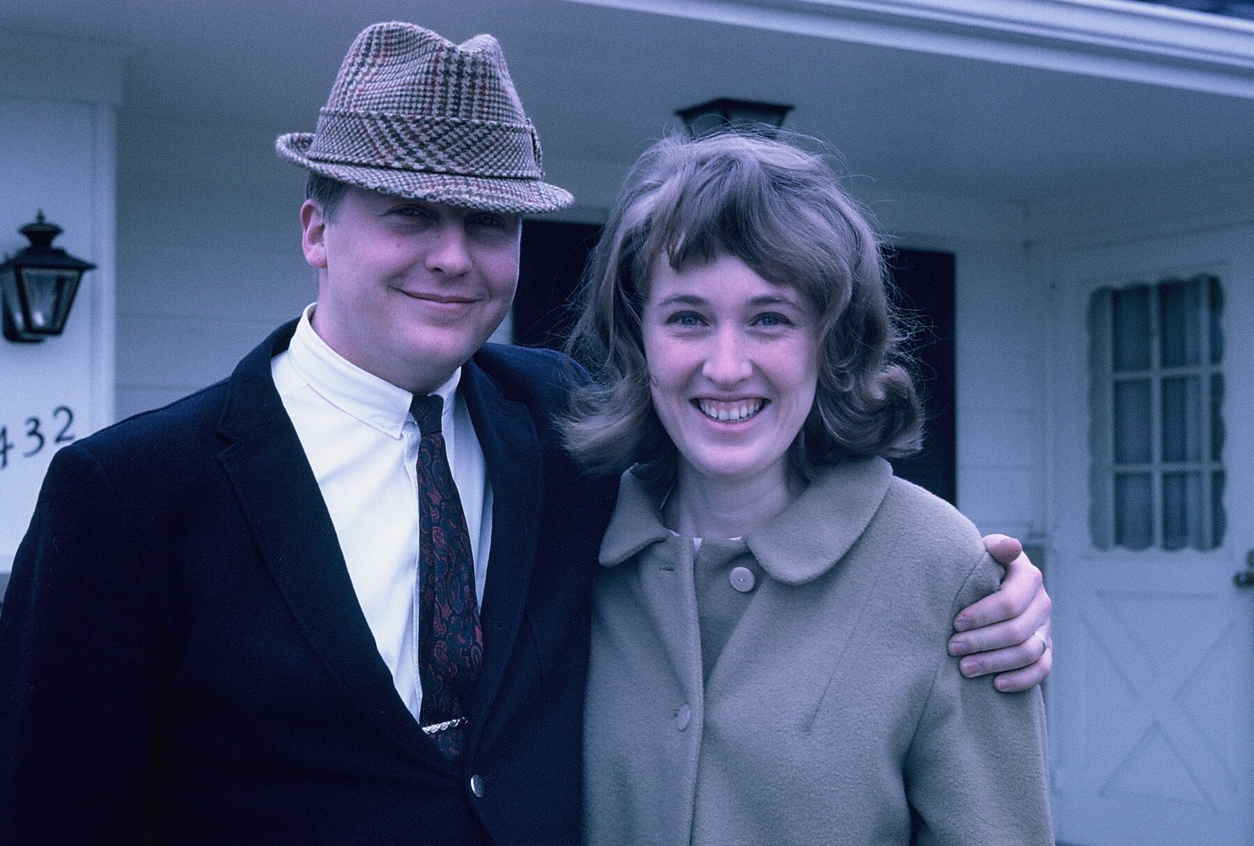 Man in a suit and fedora with his arm around a smiling woman in a beige coat, outside a house.