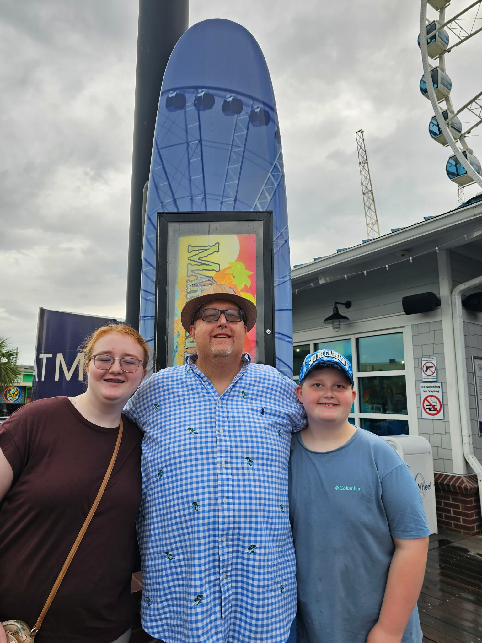 Three people posing in front of a tall blue surfboard sculpture at an outdoor amusement area, with a ferris wheel reflection on its surface.
