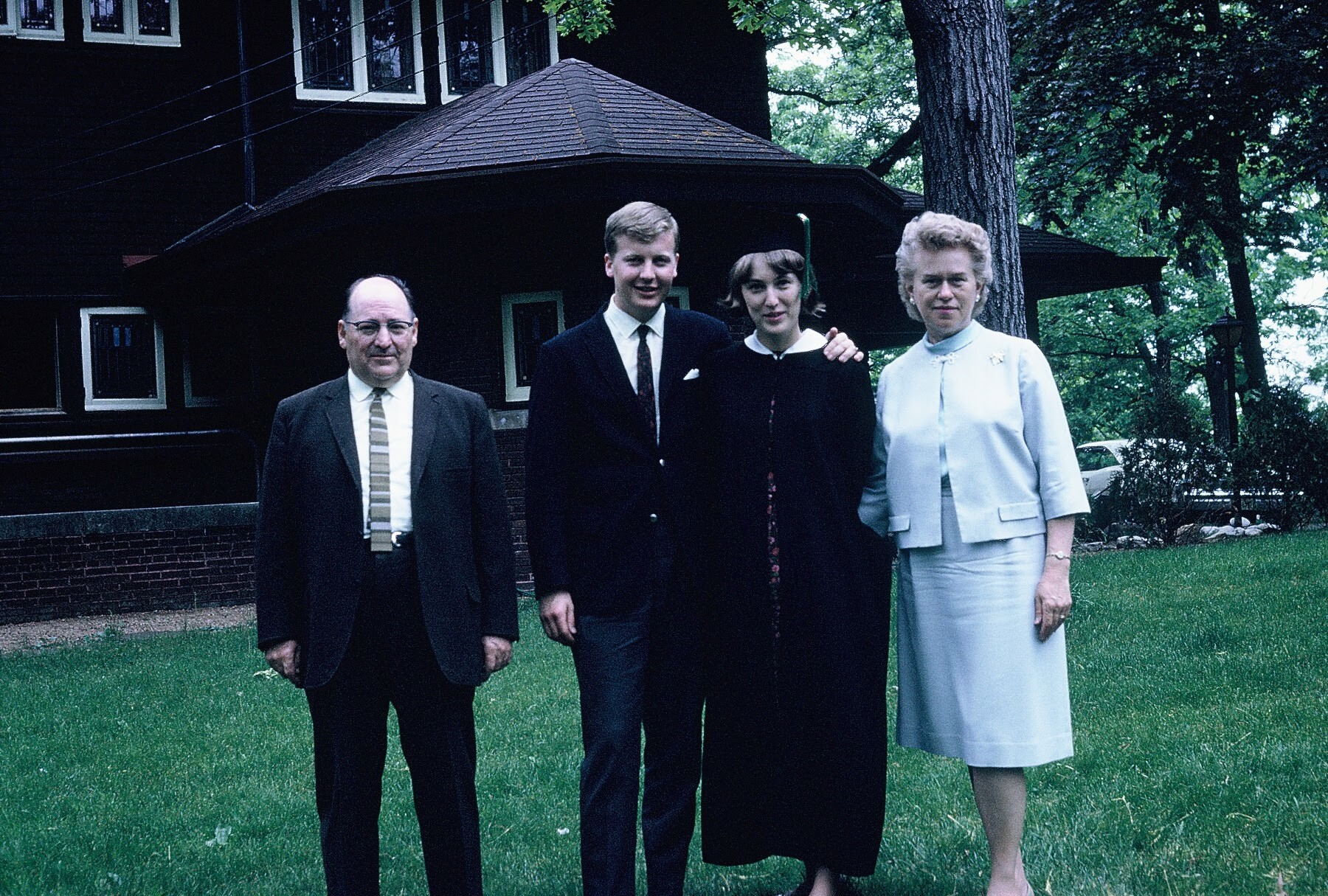 Group of four adults posing outdoors on a grassy lawn in front of a dark wooden house, all dressed in formal attire (two men in suits, two women in dresses).