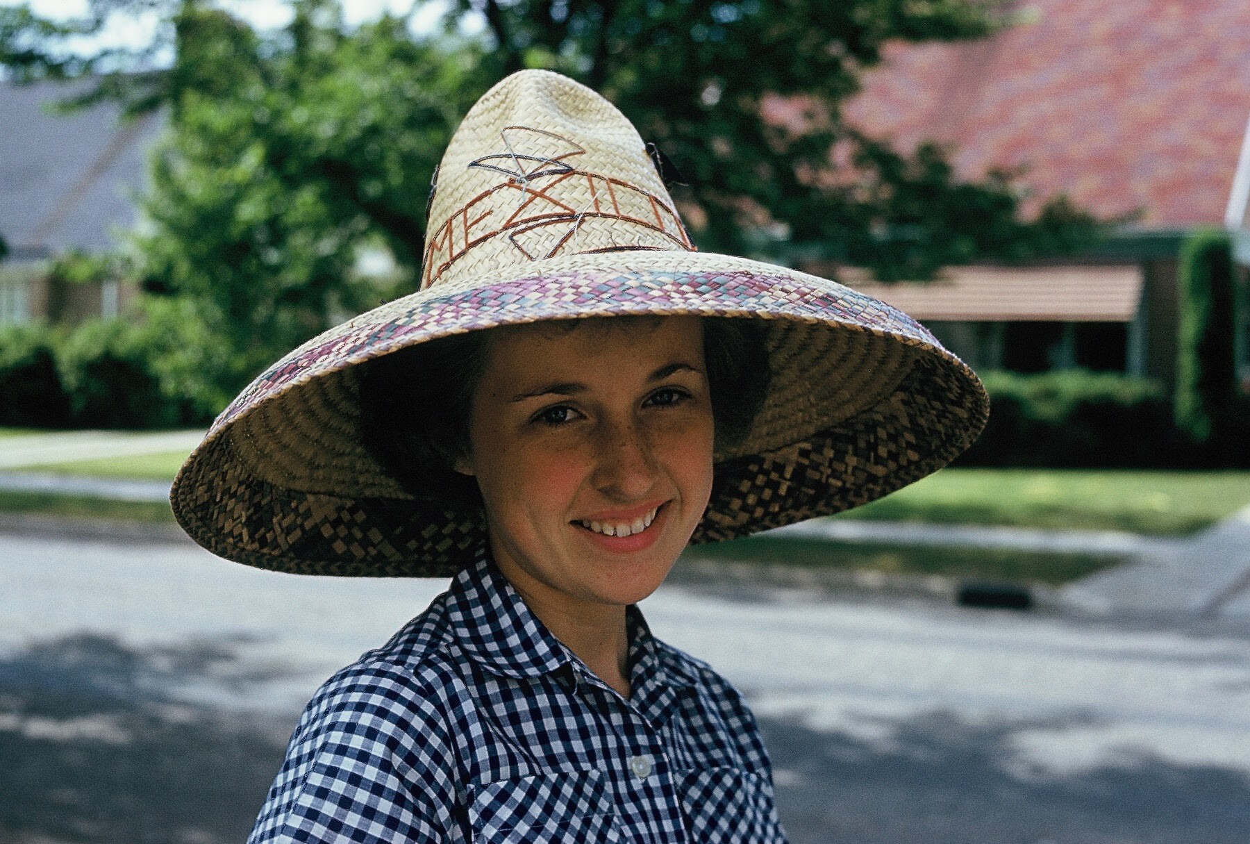 Smiling young girl wearing a large straw sun hat with decorative stitching, outdoors on a sunny day.