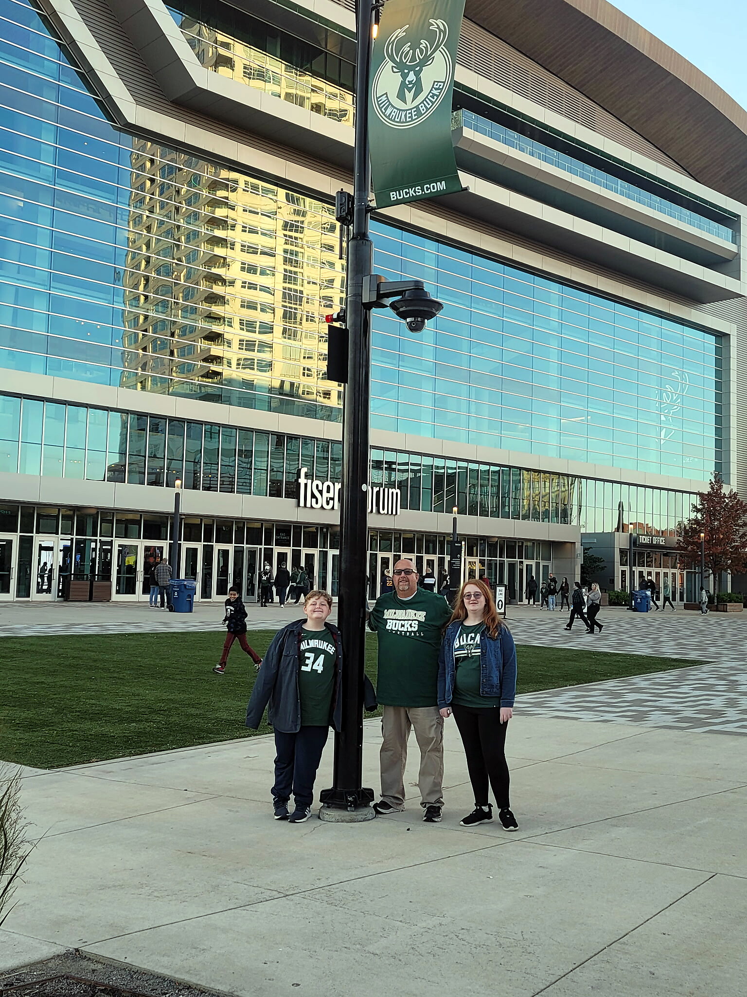 Three people in Bucks shirts pose together in front of a black lamppost at Fiserv Forum stadium exterior.