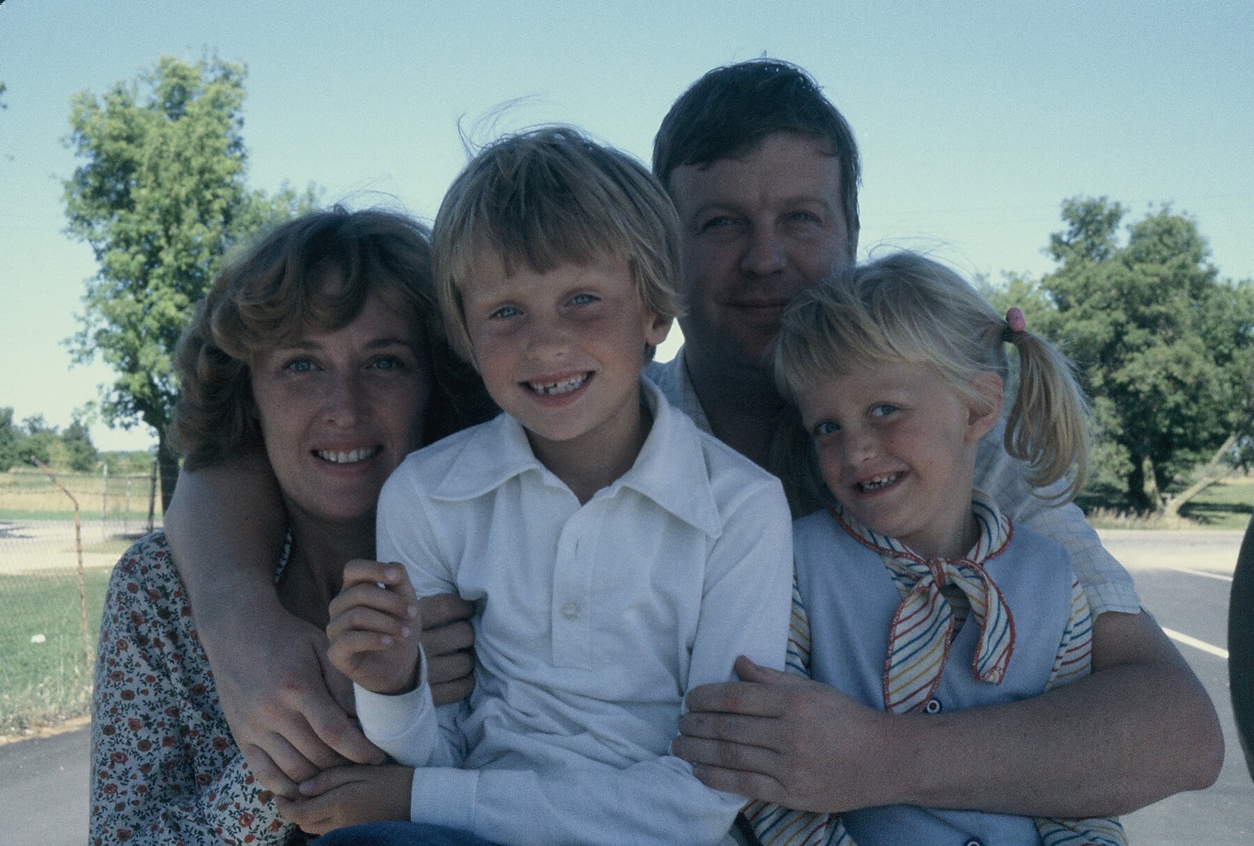 Family of four posing together outdoors, smiling at the camera with arms around each other.