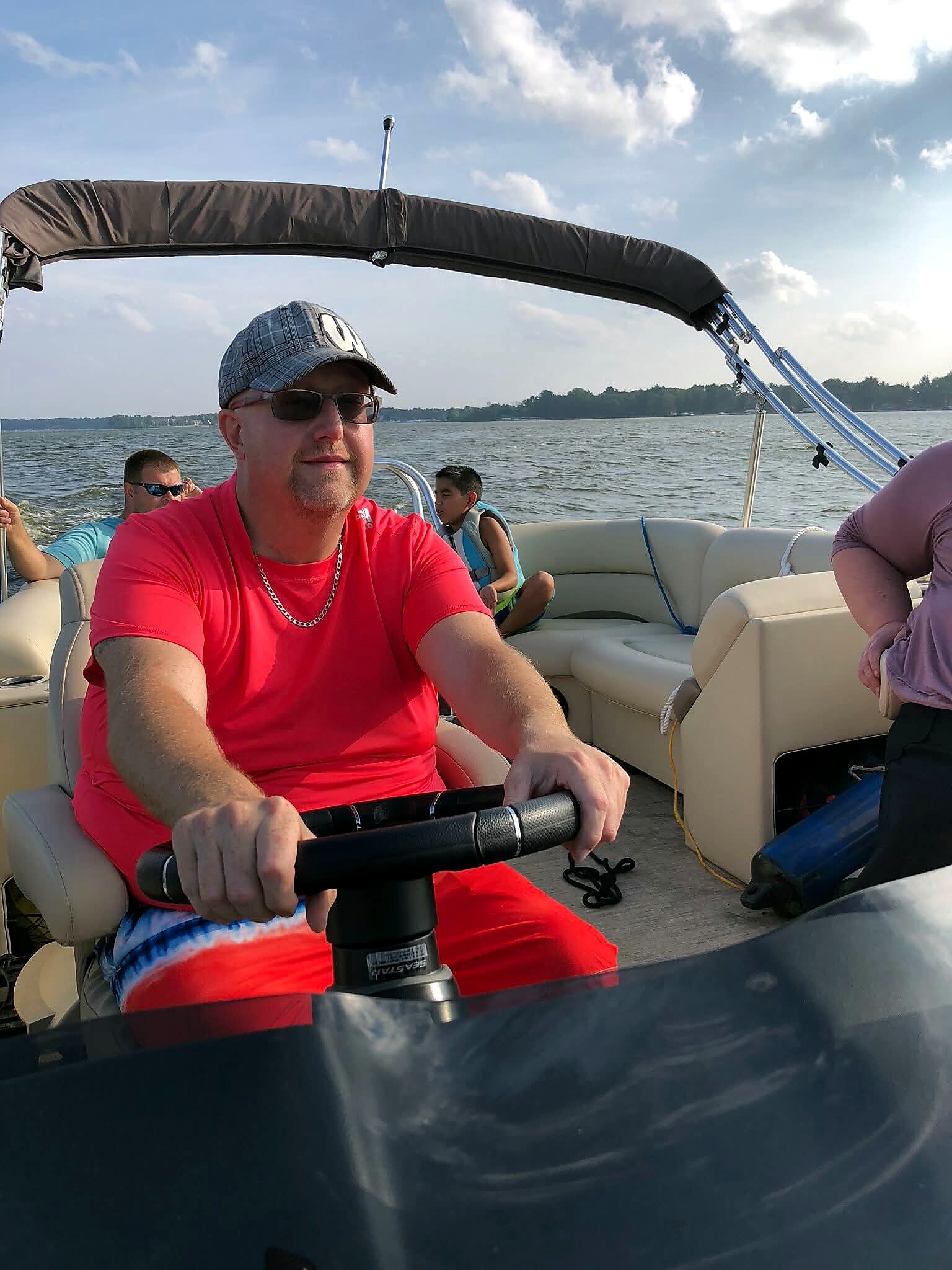 Man in a bright red shirt steering a speedboat, with two passengers and open water behind her/him.