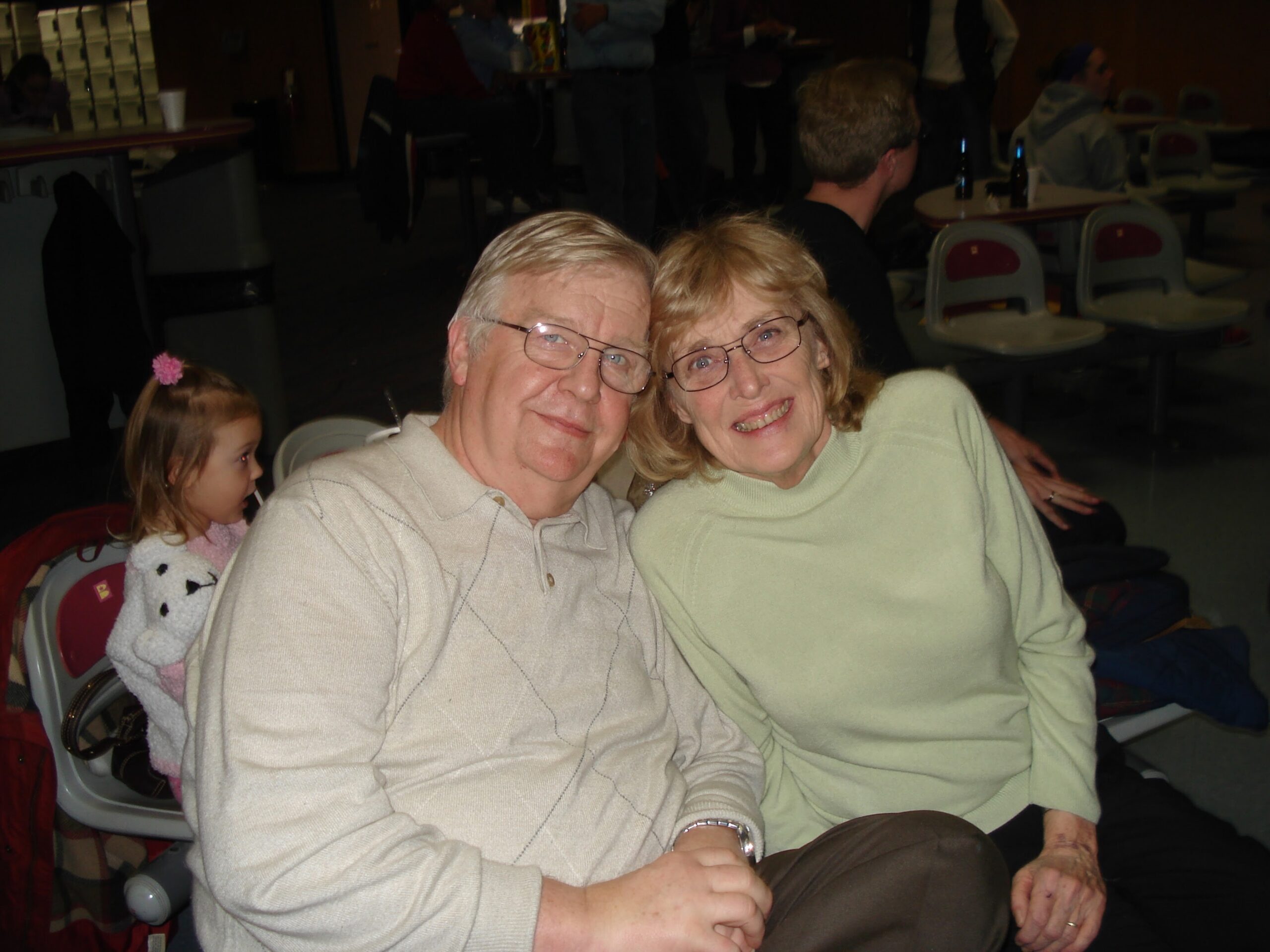 Smiling elderly couple seated together at a casual indoor gathering, wearing light sweaters.