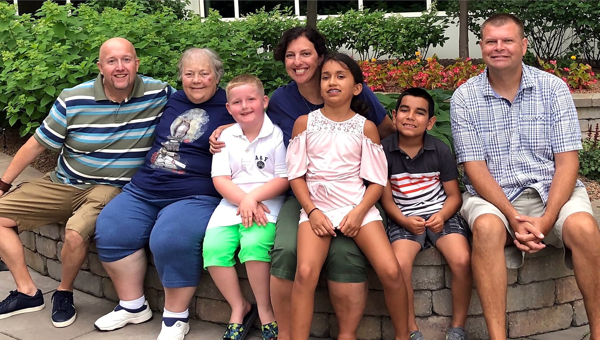 Seven people sit on a stone wall outdoors, smiling, with green bushes and colorful flowers behind them.