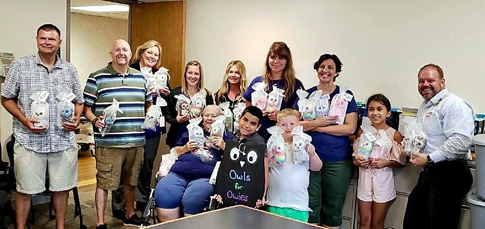 Group of adults and children posing in an office, each holding gift bags for a charity fundraiser with a chalkboard sign nearby reading 'Owls for Owies'.