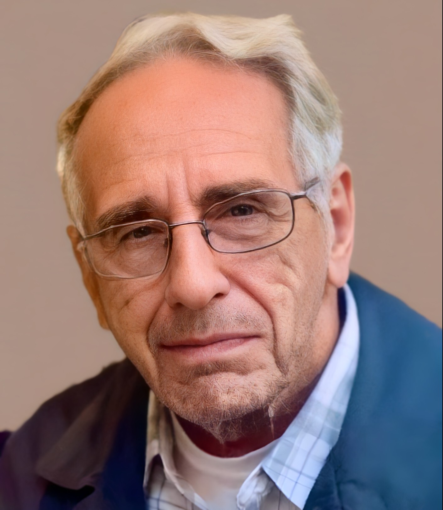 Close-up portrait of an older man with gray hair and glasses, wearing a blue jacket and checkered shirt, looking slightly off-camera.