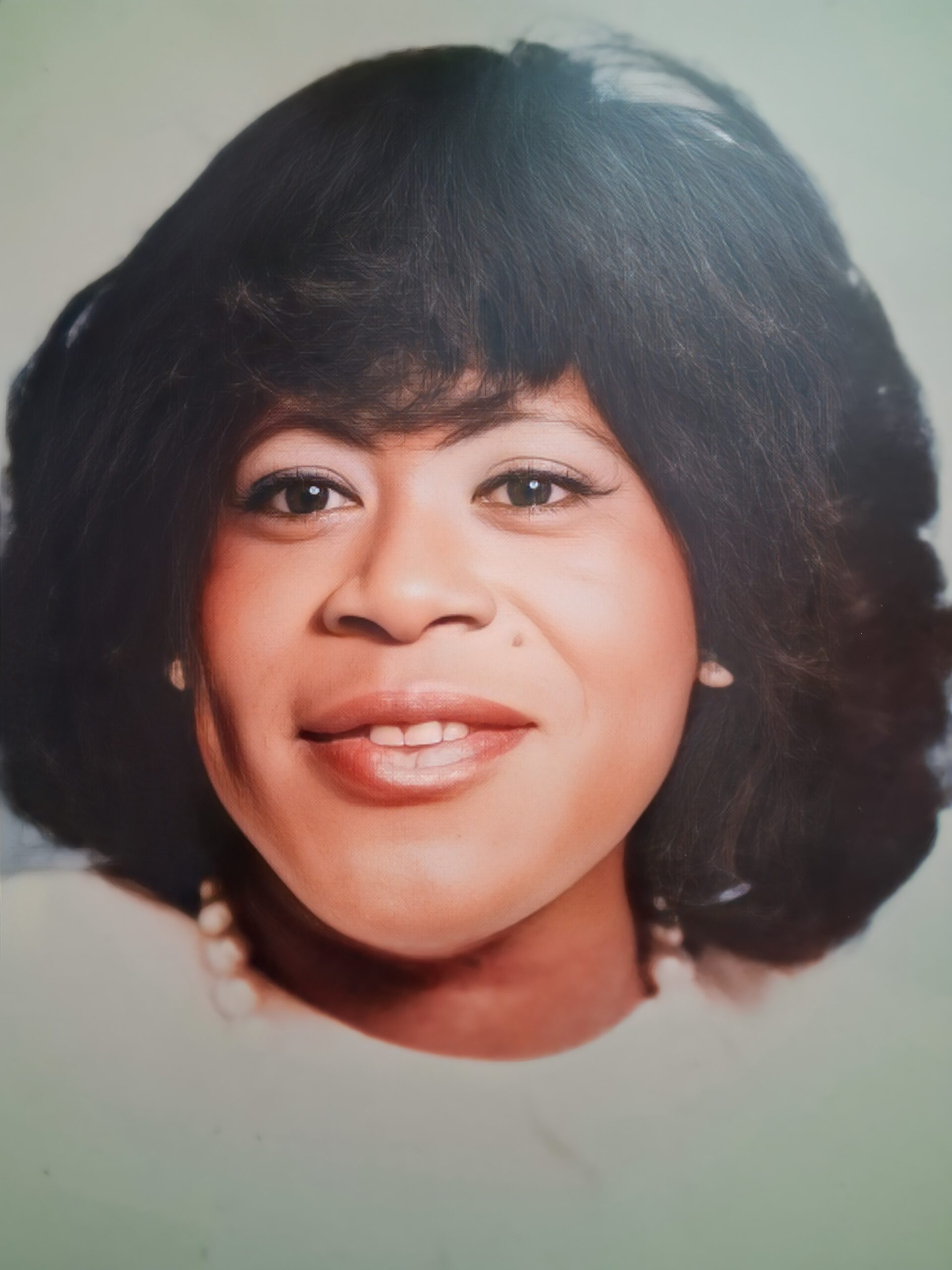 Close-up portrait of a smiling woman with dark hair and a light background, wearing a pearl necklace.