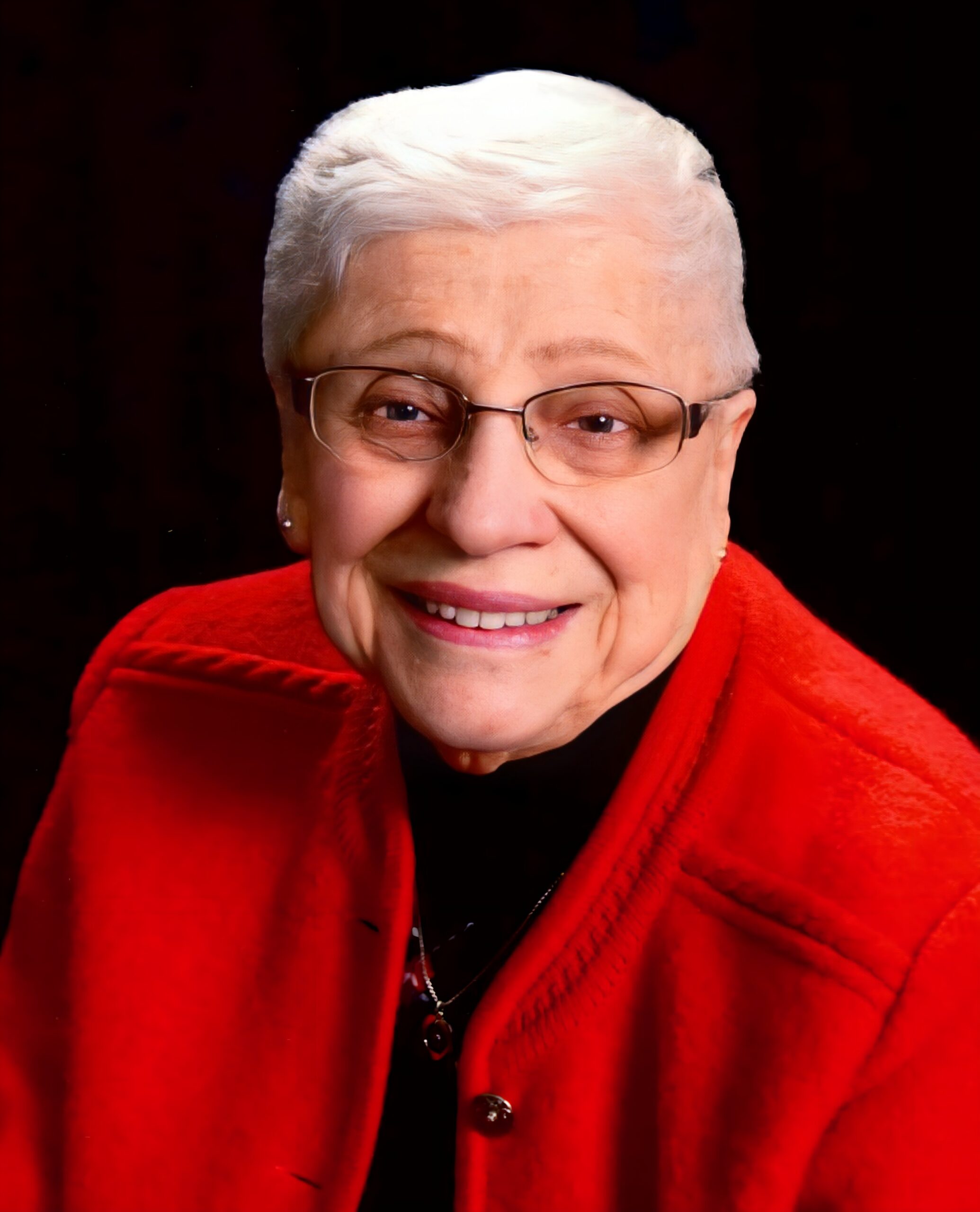 Close-up of a smiling elderly woman with short white hair, glasses, and a red coat against a dark background.