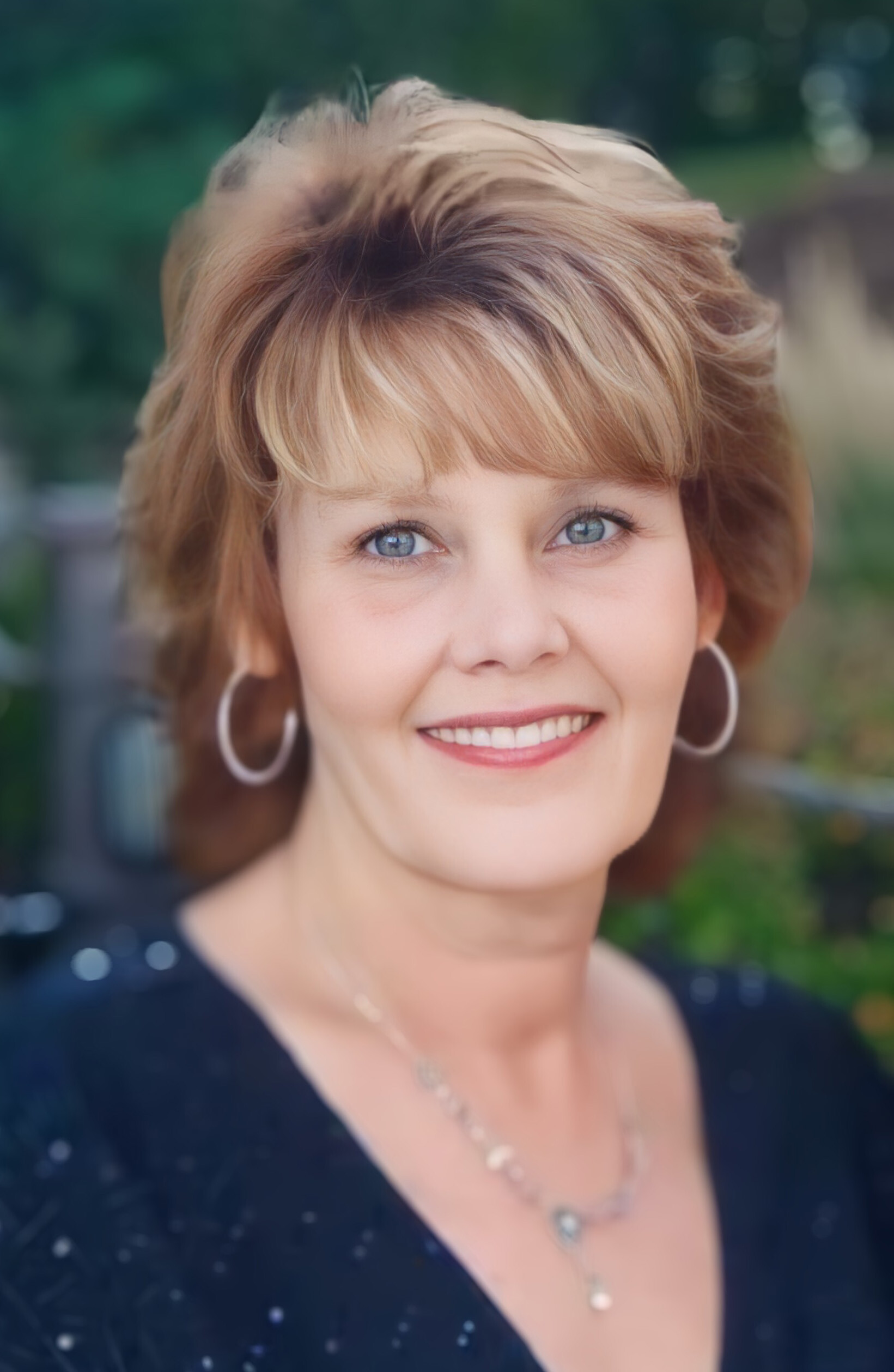 Close-up portrait of a smiling woman with light brown hair and hoop earrings, outdoors with blurred greenery in the background.