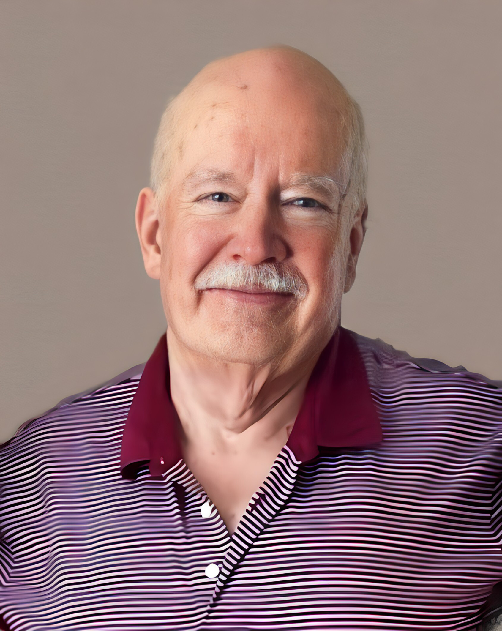 Close-up portrait of an smiling older man with gray mustache, wearing a maroon and white striped polo shirt against a neutral background.