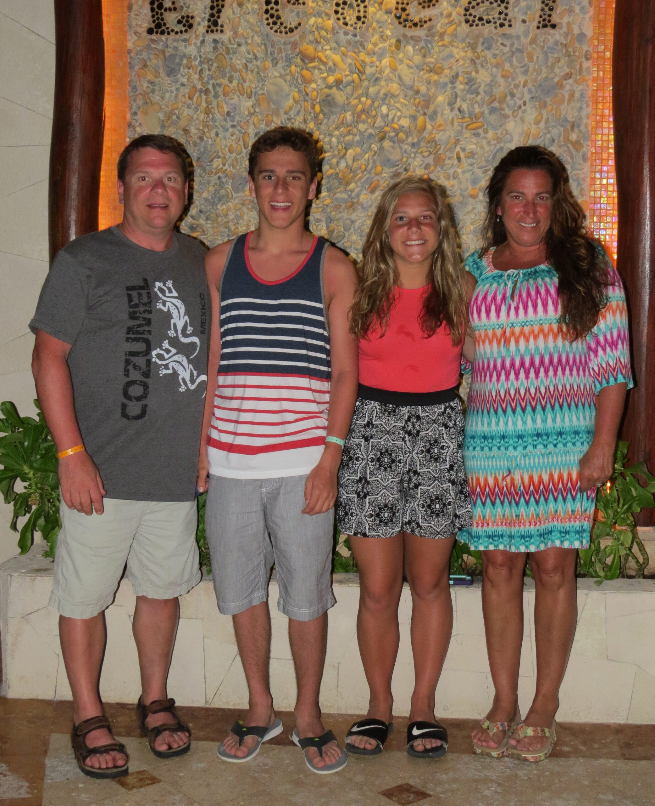 Four people posing together indoors in front of a pebble mosaic wall, wearing casual summer outfits and sandals, smiling.