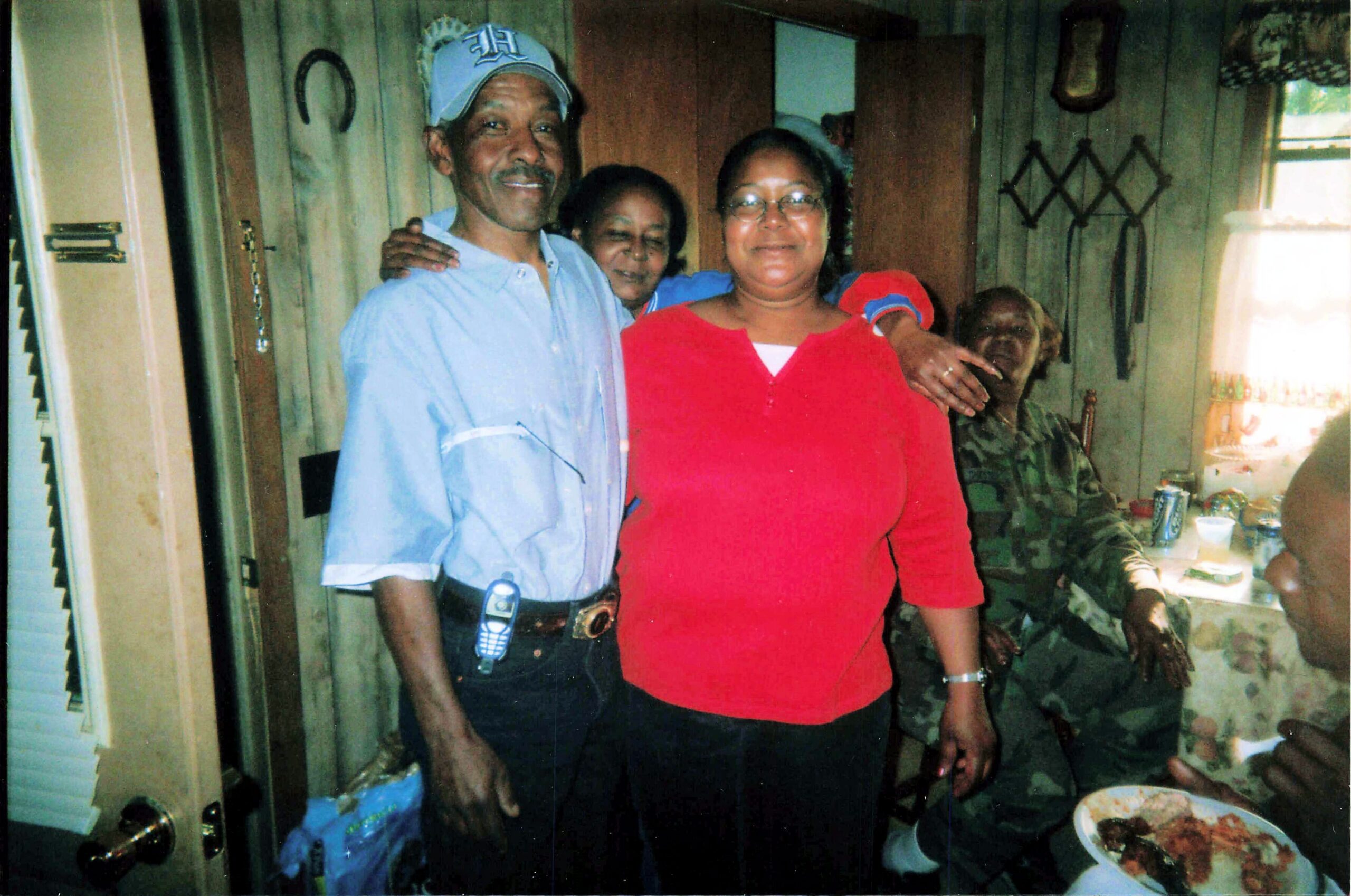 Man in a blue shirt and cap with his arm around a woman in a red top, posing with others inside a home; food and drinks on the table to the right.