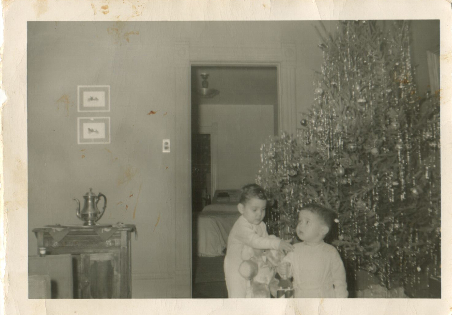Two young children near a decorated Christmas tree in a cozy living room; one holds a stuffed animal while the other watches nearby.