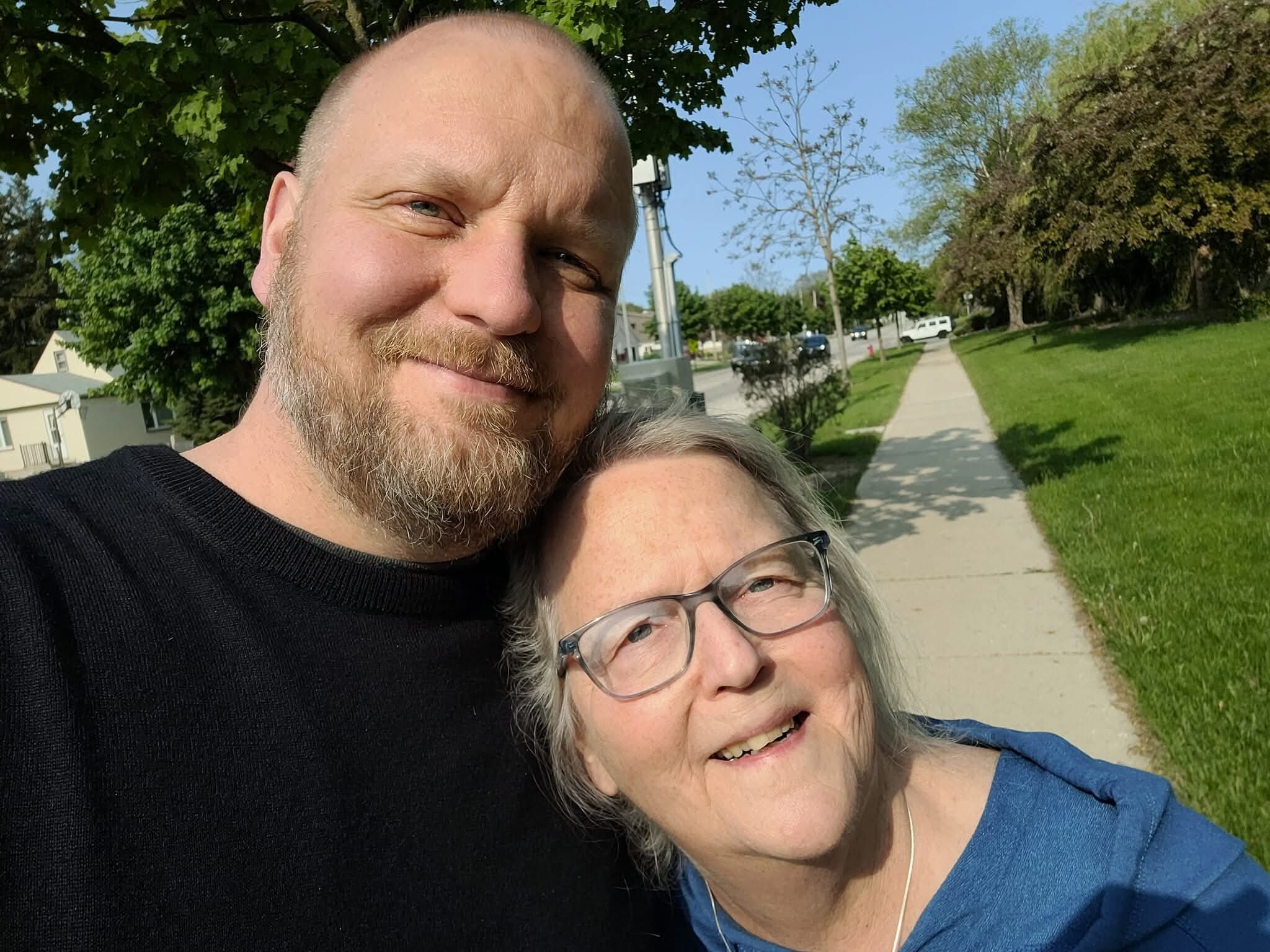 Close-up of a smiling couple taking a selfie outdoors on a sunny day, with a sidewalk and green trees in the background on a residential street.