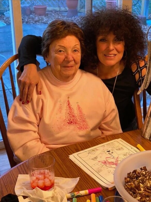 Two women posing indoors at a dining table; elderly woman in a pink sweater sits with a younger woman who has curly hair.