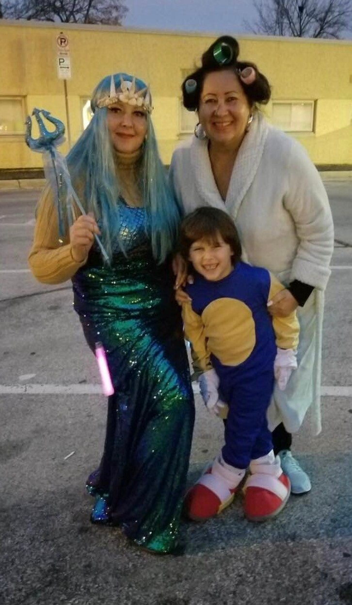 Three people in costumes posing outdoors: a mermaid in a teal sequined gown, a woman with curlers, and a smiling child in a blue and tan animal onesie/suit.
