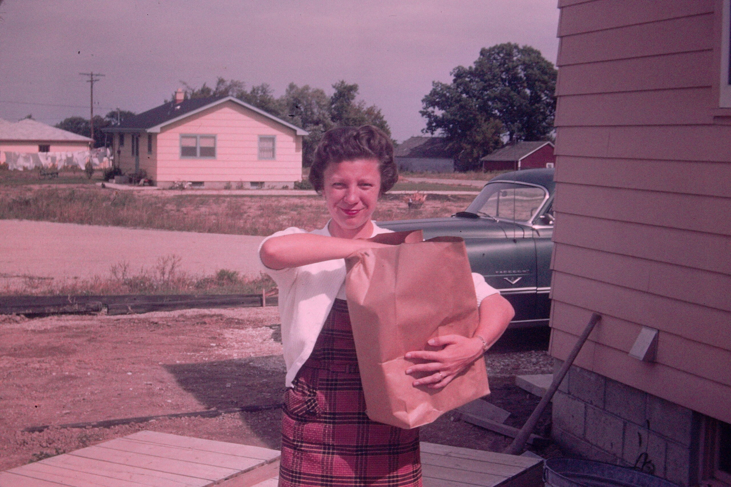 Woman standing outside, holding a brown paper grocery bag in a suburban yard.