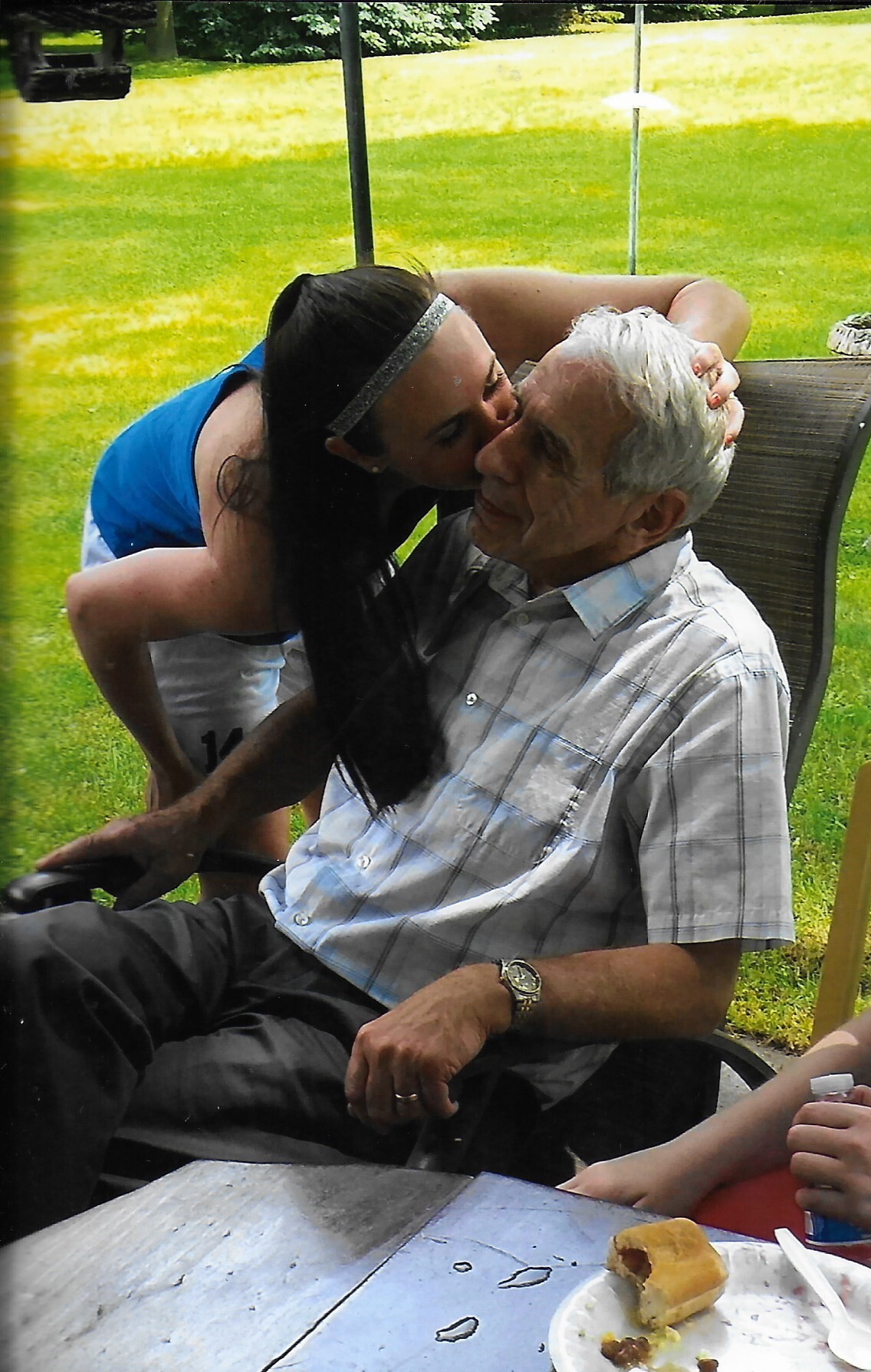 Young woman kissing an elderly man in a wheelchair outdoors, at a picnic table.