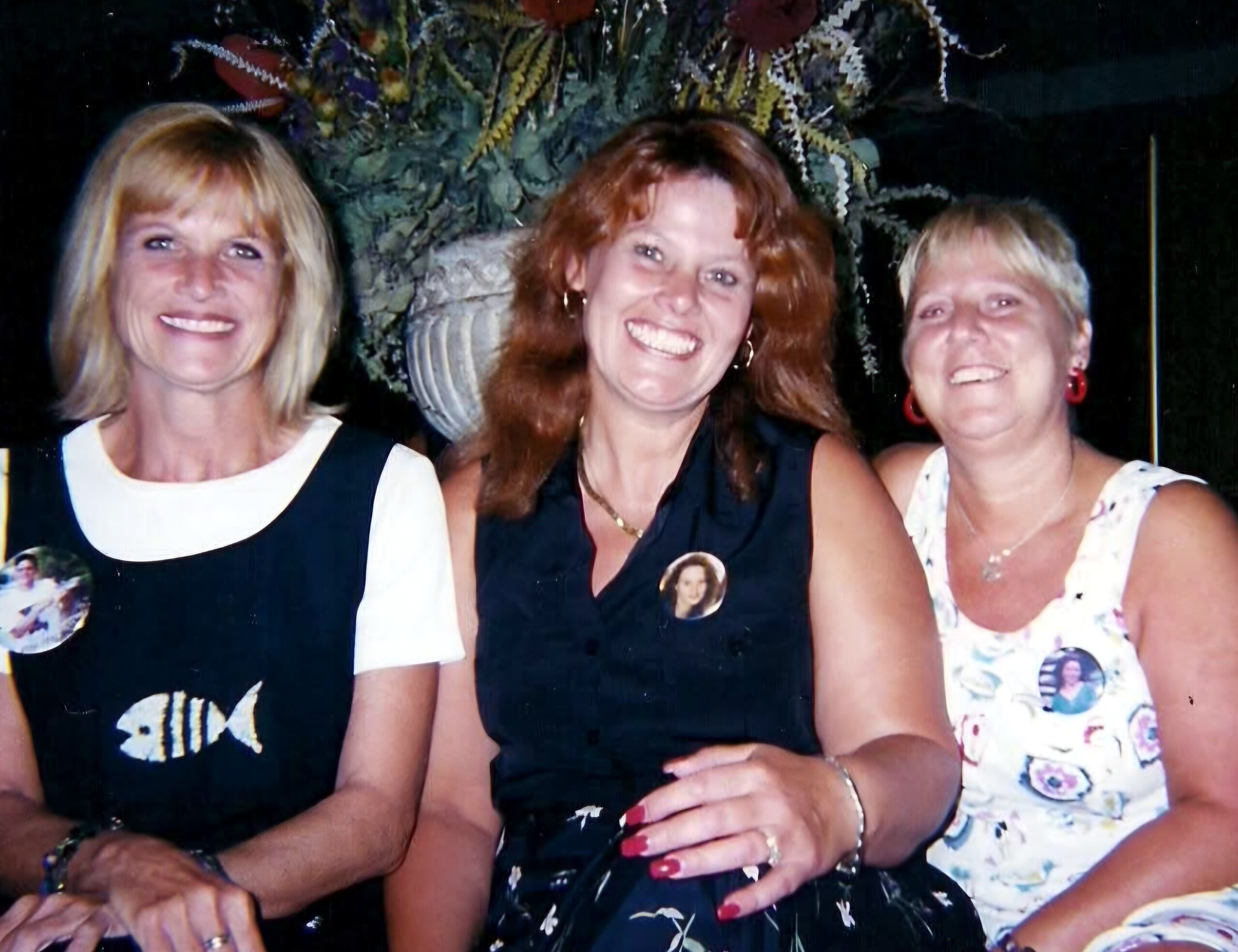Three women posing closely together, smiling for a group photo at night, wearing dresses with round badges.