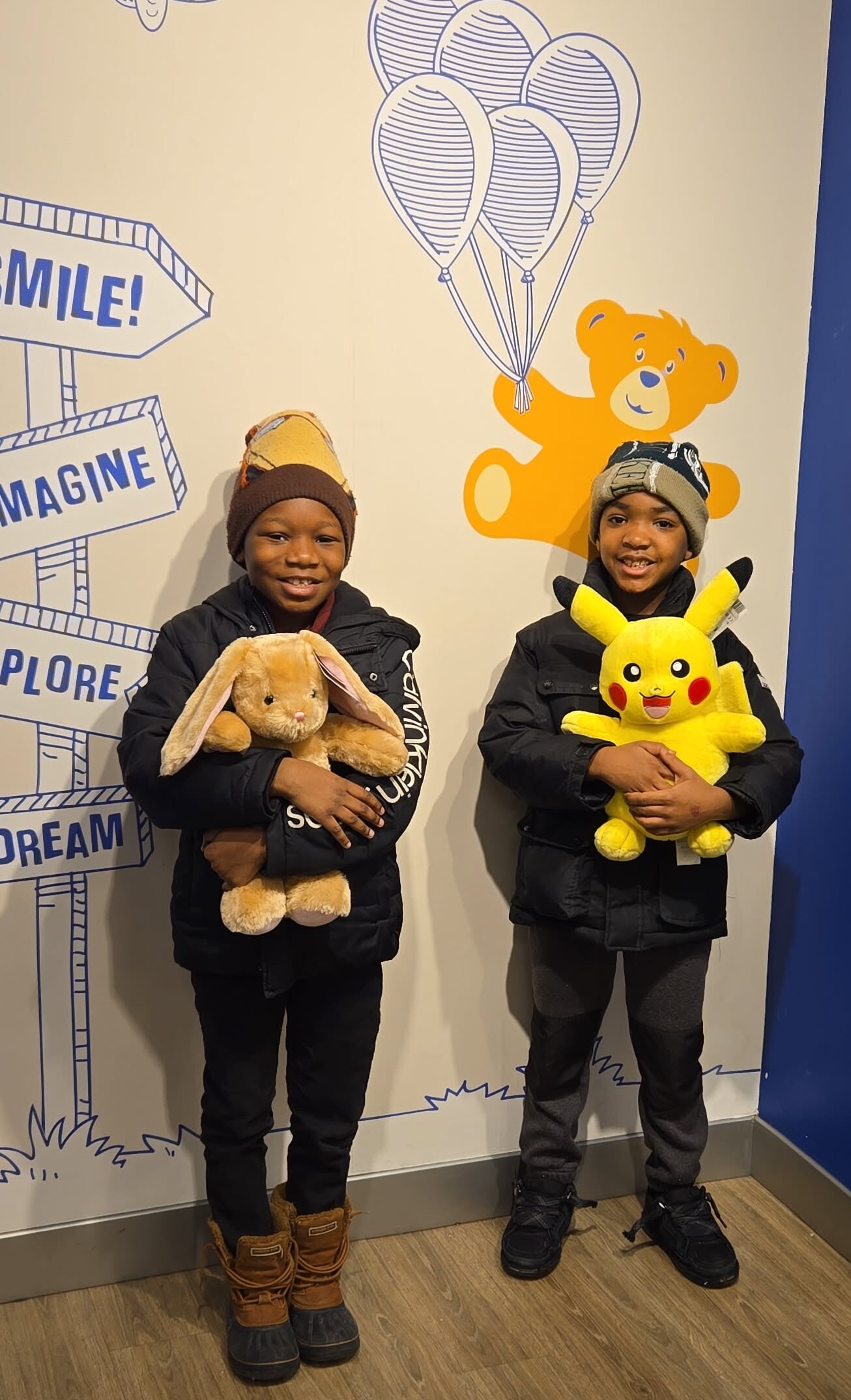 Two smiling children in winter jackets and hats hold plush toys—a bunny and Pikachu—standing in front of a mural with balloons and signs.